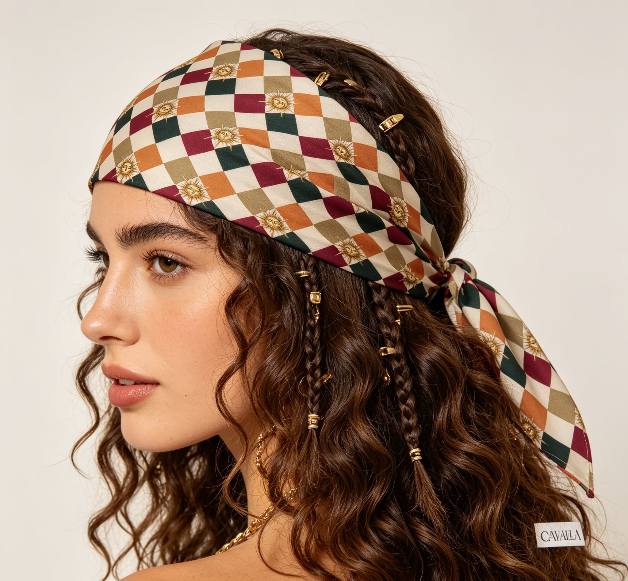 A woman with long, wavy brown hair models a patterned headscarf. The scarf features a multicolor harlequin diamond pattern in cream, rust orange, burgundy, and olive green, accented with small gold medallions and gold-toned hair ties and beads.
